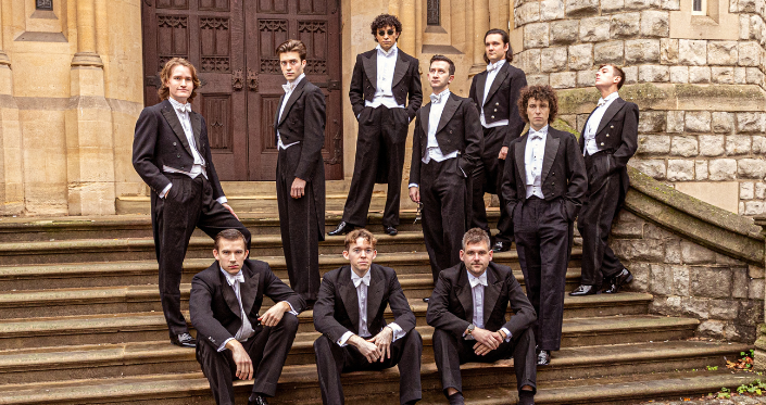 10 young men in smart suits, posing on the steps of a historical building.